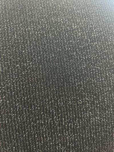 The image is a close-up, abstract view of a textured surface. The surface appears to be a dark, charcoal gray material with a repeating pattern of small, raised bumps or indentations. Interspersed among these darker elements are numerous small, bright white or silver flecks that catch the light, creating a shimmering or sparkly effect. The texture is uniform across the frame, with no distinct objects, people, or a clear scene visible. The lighting highlights the texture, with some areas appearing brighter where the flecks are more densely packed or catch the light at a certain angle. There is no discernible time of day, weather, location cues, or emotions conveyed by this image. No text is visible. The overall impression is of a material with a finely textured, somewhat iridescent surface.