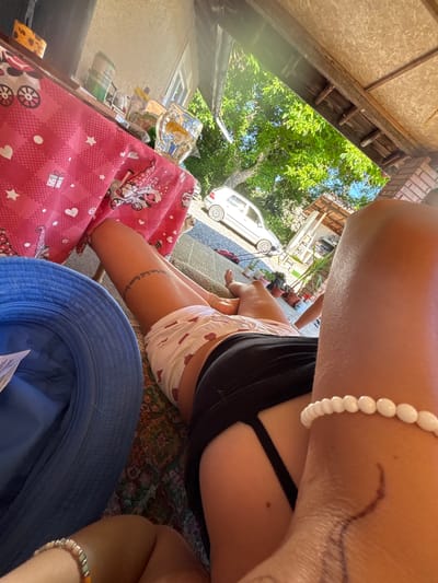 The image captures a sunlit outdoor scene, likely a backyard or patio area, with a person lying down. A blue bucket hat is placed on the left side of the frame. In the center, a person's legs are visible, wearing patterned shorts and a dark bikini bottom. A tattoo is visible on their left thigh. To the right, another person's arm and shoulder are in the foreground, adorned with a white beaded bracelet. A table with a pink tablecloth featuring white stars and cartoon gnomes is in the background. On the table, there are drinks and various items. A white car is parked in the driveway in the distance. The environment appears to be a residential area with trees and a partial view of a building. The location is identified as Belopoptsi, Bulgaria.
