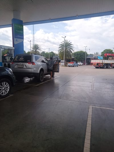 The image captures a scene at a gas station in Tha Ruea, Thailand. A silver Mitsubishi Pajero Sport SUV, license plate ทพ 4925, is parked at pump number 4, adjacent to a blue and white sign advertising "Gasohol E20", "Gasohol 91", and "ดีเซล Diesel." A person stands by the driver's side door of the SUV, facing into the vehicle. Another vehicle is partially visible in the foreground, positioned to the left of the SUV. In the background, there is a blue pickup truck, a white pickup truck, a white van, and a red and white crane truck. The scene is set on a concrete surface, likely the gas station forecourt, under a partially cloudy sky.