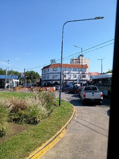 The image shows a street in San Fernando, Argentina (latitude: -34.435, longitude: -58.567).  A gray pickup truck is parked on the right side of the street.  The truck is facing away from the camera. The pickup truck is on a paved road with a yellow line down the center. The road curves to the left and there is green grass and bushes growing on the outside of the curve. A street lamp is visible on the left side of the image.  A building, with a red-tile roof, is visible in the background. There is a sign on the building that says "Hotel". The sky is blue with some white clouds.
