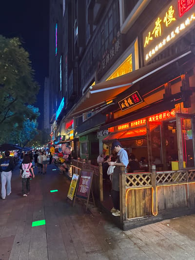 This image depicts a lively street scene at night in Longta, China. The foreground is dominated by a wooden sidewalk with glowing green markers embedded in it, suggesting a designated walking path or perhaps an interactive element. Several people are walking along this path, some dressed casually.
To the left of the sidewalk, a building with a traditional wooden facade is visible, adorned with warm, yellow neon lights outlining its structure and a second-story balcony. Inside this building, a few patrons can be seen dining or socializing. Above the entrance, a prominent neon sign displays Chinese characters. Further down the street, more shops and buildings are illuminated by various neon signs, creating a vibrant and colorful atmosphere.
In the mid-ground, a group of people are gathered around a vendor selling balloons. The colorful balloons, including some shaped like cartoon characters, add a festive touch to the scene. The overall mood appears to be one of enjoyment and relaxation, typical of a bustling night market or entertainment district.
The text visible in the image includes:
- "从前慢两周年店庆" (Congqian Man two-year anniversary celebration)
- "全体按摩" (Full massage)
- "从前慢" (Congqian Man) - repeated several times, likely the name of a business.
- "在有她的地步通是有趣的人" (In the place where she is, everyone is an interesting person)
- "《林里》柠檬首先要快乐 其次都是其次遇见你很开心呀" (《Linli》 Lemon, first be happy, then everything else is secondary. Meeting you makes me very happy!)
The time of day is clearly night, as indicated by the darkness of the sky and the extensive use of artificial lighting from neon signs and building interiors. The presence of trees suggests an urban park or tree-lined street. The weather appears to be clear, allowing for outdoor activities.