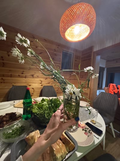 A close-up shot shows a woman's hand holding a glass vase filled with white daisies. The vase is on a white circular table set for a meal. The table is laden with food, including a bowl of green salad, a platter of dark meat, and a dish of sliced bread. A green bottle of drink and a glass of amber liquid are also visible. In the background, the room is paneled with light-colored wood, and a woven pendant lamp hangs from the ceiling. Two chairs are visible, and a doorway leads to another room. The scene appears to be set in a home or restaurant in Virak, Montenegro.
