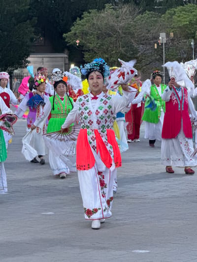 A vertical, eye-level outdoor shot captures a procession of women in brightly colored traditional Hanfu clothing in Jinzhou, China. The foreground features the participants walking on a gray concrete pavement. Lush greenery and a portion of a building rise in the background, adding depth to the scene. The women's garments, adorned with vivid hues and intricate patterns, contrast sharply with the neutral background. The clothing incorporates reds, greens, blues, and various other shades, contributing to the overall visual interest.