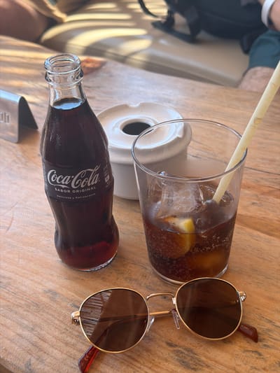 A Coca-Cola bottle and a glass of the beverage with ice and a slice of lemon sit on a wooden table. A pair of sunglasses rests in the foreground. In the background, partially visible, are two people sitting on a couch. The scene appears to be an outdoor seating area of a cafe or restaurant in Sant Josep de sa Talaia, Spain.