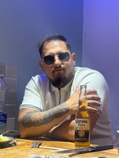 A man with a beard and sunglasses sits at a wooden table. He wears a white polo shirt with black stripes on the collar and sleeves. His left arm is bent, and he rests his hand on his bicep. A bottle of Corona Extra beer stands on the table in front of him, positioned between his chest and the viewer. To the left of the beer bottle, a partially visible bottle of "Aqua Pura" water and a grey brick wall can be seen. The man has tattoos on his left arm. The background is a gradient of blue and purple, suggesting artificial lighting or a colored wall. There is a fork, a knife, and some lime wedges on the table. The scene appears to be indoors.