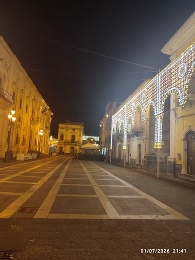 The image captures a nighttime scene in the city of Militello in Val di Catania, Italy. The foreground features a wide, paved piazza with a geometric pattern of light and dark square tiles intersected by light-colored strips. A storm drain is visible in the lower left corner of the piazza. The piazza is illuminated by streetlights and decorative lights adorning the surrounding buildings.
On the left side of the piazza, a large, ornate yellow building stands, its facade decorated with architectural details and balconies. Streetlights with multiple lamps are positioned along the sidewalk in front of this building. A banner with Italian and European flags is attached to the facade of this building.
To the right of the piazza, another building is visible, also illuminated. Further down the piazza, a structure with a yellow canopy is present. The right side of the image is dominated by a long, historic building extensively decorated with intricate patterns of white and yellow string lights, creating an archway effect. These lights highlight the building's architectural features, including arched windows and balconies.
In the background, additional buildings and a darker sky complete the scene. The overall impression is of a public square decorated for a festive occasion. The date stamp "01/07/2026 21:41" is present in the lower right corner of the image.