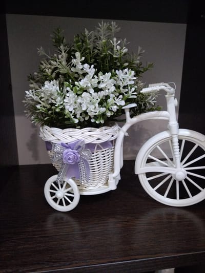 A white decorative bicycle basket filled with artificial white flowers and greenery sits on a dark wooden surface. The bicycle has three wheels, the largest at the front, and a handlebar. A purple ribbon with a silver rose adorns the basket. The background is a dark, indistinct space.