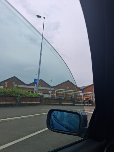 The image depicts an overcast daytime scene from the interior of a vehicle, looking out through a partially lowered, tinted driver's side window towards a retail park in Stafford, United Kingdom.
In the immediate foreground, the dark blue or black interior trim of the vehicle's door frame is visible on the right, bordering the curved edge of the car window. A blue-tinted side mirror, positioned on the lower right, reflects a segment of a blue vehicle, likely the one from which the photograph was taken. Below the mirror, a section of asphalt road is visible, marked with a solid white line and bordered by a light grey concrete curb.
The midground features a low brick wall, constructed from reddish-brown bricks, topped with dark capstones and overgrown with dense green foliage. Behind this wall, a tall, slender grey metal streetlight pole ascends, capped with a dark grey, angular light fixture. Attached to this pole, a prominent rectangular blue sign displays a white international symbol of access (wheelchair user).
Behind the streetlight and wall, a large, red-brick building with multiple gabled roofs dominates the scene. The building, identified as a Sainsbury's supermarket from visible signage in crops, features at least three distinct gabled sections with circular decorative elements or windows on their facades. Below the gables, a continuous white framework or architectural detailing spans the building. Additional blue signs with the white international symbol of access are visible closer to the building's entrance areas. A yellow triangular sign with partially obscured black text, reading "ENTRY SLOW DOWN," is also visible near the building.
The background consists of a uniformly grey, heavily overcast sky. No individuals are visible within the frame.