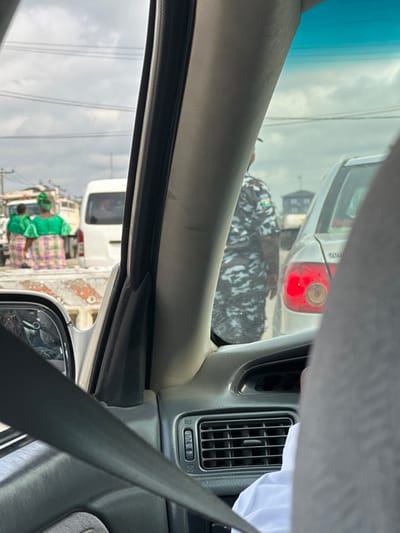 The scene depicts a street view in Eneka, Nigeria, observed from inside a vehicle, likely stationary in traffic. The foreground reveals a gray car interior, including a segment of the dashboard with an air vent, a partial view of a black seatbelt traversing diagonally, and a white-sleeved arm of an occupant. Through the windshield, a congested roadway is visible under an overcast sky.
In the midground, a silver sedan is stopped ahead, its left rear brake light illuminated. To the right of this sedan, a person wearing a blue, white, and black camouflage uniform, indicative of a security or police officer with a visible badge on the left sleeve, stands facing away from the viewer. To the left, a white van is present in traffic, followed by a flatbed truck. On the rear of the flatbed truck, two individuals, likely women, are visible, wearing matching vibrant green blouses with puffed sleeves and colorful patterned wrapper skirts and head coverings. One individual appears to be holding a small child, also in green attire. Utility poles and power lines are discernible in the background against the sky. A concrete road barrier is partially visible near the car's side mirror. No direct interactions between individuals are discernible.