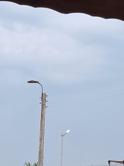 The media file depicts an outdoor scene, likely in Bulgaria, with a pale blue sky as the dominant background. The sky suggests a daytime setting, possibly overcast or a hazy sunny day.
The primary subjects are utility poles and their associated infrastructure. One long, wooden pole stretches horizontally across the frame, from left to right, supported by wires that are visible against the sky. Several small, dark objects, possibly insulators or birds, are attached to this pole. A streetlight is mounted on the end of this pole, hanging downwards. Further to the right and higher up, another streetlight is visible on a separate pole, illuminated and pointed towards the viewer. There are also several thin, dark wires crisscrossing the sky, connecting to the poles.
On the far left edge of the image, a dark, curved object is partially visible, obscuring a portion of the sky. Its texture suggests it could be a building overhang or a natural element like a tree branch. In the lower right, some green foliage is present, hinting at vegetation in the vicinity.
No people are visible in the frame, nor are any specific events taking place beyond the static presence of the utility infrastructure. The scene is likely a view of a street or an area with overhead power and lighting. The overall impression is one of everyday infrastructure against a broad sky. There is no visible text in the image.