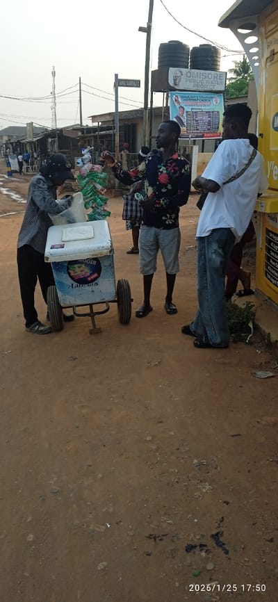 A street scene shows a vendor with an ice cream cart serving customers. One customer wears a floral shirt and shorts, while another wears a white shirt and jeans. A sign for the “Omisore Public Water Project” is visible in the background, along with a poster for a Cherubim & Seraphim church. The scene is captured on a dirt road with buildings in the distance. A MTN-branded kiosk can be seen in the background. The date stamp indicates “2026/1/25 17:50.”