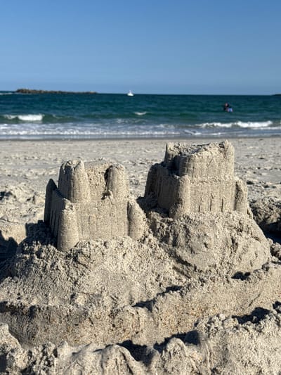 This is a close-up, eye-level shot of two sandcastles on a sandy beach. The sandcastles are the primary focus, with detailed textures and crenellated tops. In the background, the ocean with moderate waves and a clear blue sky are visible. A distant rocky outcrop is on the left, and a small white sailboat and two people in the water are on the right. The scene is bathed in sunlight, casting shadows on the sand. The location is identified as Wrightsville Beach, United States of America.