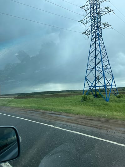 The image depicts a roadside scene near Кокино, Russia under an overcast sky. A blue high-voltage transmission tower stands in a field of green grass beside a paved road with a white line marking its edge. The road surface appears wet, suggesting recent rain. The side mirror of a vehicle is visible in the lower right corner, indicating the picture was taken from inside a car. The background shows a line of trees, further into the landscape.
