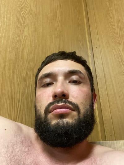 A close-up, chest-up, selfie-style photograph of a man with a full beard and dark hair is presented. He is facing the camera directly, with his head angled slightly down. The background consists of a light brown, wooden paneled wall with vertical lines. The man's skin appears reddish, potentially from sun exposure or flush. His eyes are open, and his lips are slightly parted. He is shirtless, with visible hair on his chest and shoulder.