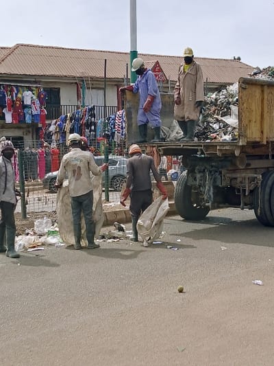 The image captures a waste collection scene on a paved street in Kwekwe, Zimbabwe. Five sanitation workers are visible, actively engaged in clearing refuse. A large, high-sided garbage truck, filled with miscellaneous trash, dominates the right side of the frame.
Two workers stand on the back of the truck. One, wearing a light blue overall, white hard hat, dark face mask, and dark gumboots, leans on the truck's left side. The other, clad in a beige overall, yellow hard hat, dark face mask, and dark gumboots, stands further back on the truck bed.
Three additional workers are at street level. One on the left wears a light long-sleeved shirt, dark trousers, dark gumboots, and a dark face mask, holding a large, light-colored refuse bag. Another, slightly to the right, wears a light-colored shirt, dark trousers, a white hard hat, and dark gumboots, also holding a similar large bag. A third worker, partially obscured by the truck on the right, wears dark brown overalls, red gloves, and dark gumboots, carrying another refuse bag.
The street is asphalt, with scattered litter and a small green, spherical object in the foreground. A curb separates the road from a sidewalk area on the left, behind which runs a metal fence. In the background, a two-story building with a reddish-brown tiled roof features numerous garments, including shirts and t-shirts, displayed on racks and hangers, indicative of a market stall or shop. A tall, white utility pole stands prominently in the center background. The sky appears overcast.