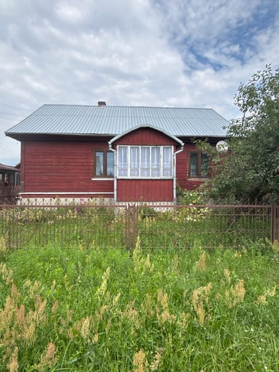A red wooden house with a metal roof is set back from the road, surrounded by a low metal fence. The house has several windows, some with white curtains visible. A small porch with windows is located in the center of the house. Tall green grass and wildflowers grow in front of the fence. The sky above is partly cloudy, with patches of blue visible. The scene is situated in Trześcianka, Poland, a village known for its rural character and traditional wooden architecture. The overall impression is one of a modest, well-maintained rural dwelling.