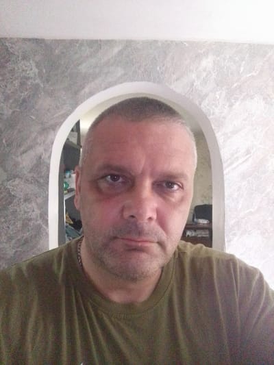 This is a close-up, selfie-style photo of a middle-aged man with short, graying hair. He is wearing a dark olive green t-shirt and a silver chain necklace. He has a serious expression, with visible dark circles under his eyes. The background features an arched doorway leading into another room. The wall on the left of the doorway has a textured, gray, marble-like pattern, while the wall on the right has a lighter, white, textured pattern. The room beyond the doorway appears to be a living space with furniture and shelves visible. The lighting suggests it is daytime, possibly indoors. There is no visible text in the image. The location context provided is Yessentuki, Russia.