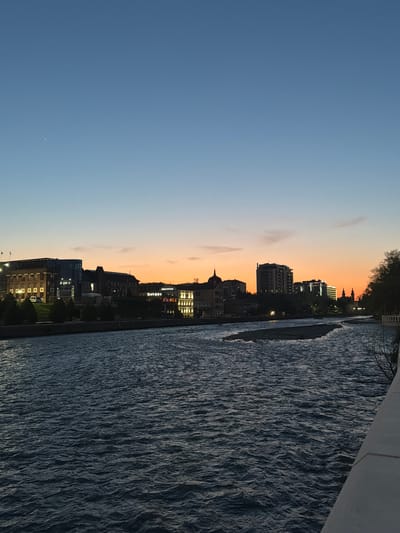 The image captures a cityscape at dusk along a river. Buildings are visible in silhouette against a gradient sky transitioning from blue to orange, with lights on in some windows. The river occupies a significant portion of the right side of the image, its surface reflecting the fading light. There is an indication of a structure on the right side that may be a bridge. The location is assumed to be Chermen, Russia based on the query. There are no people visible in the image.