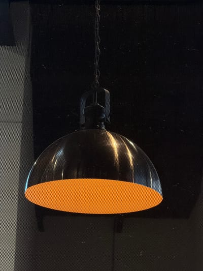 A single dark metallic, dome-shaped pendant lamp is centrally suspended by a black link chain. The lamp's exterior has a reflective finish, showing vertical light streaks. The underside of the lamp emits a bright orange illumination with a visible fine, consistent diamond mesh pattern across its surface. The background consists of a dark, uniform surface entirely covered by a subtle, fine diamond mesh texture. A vertical lighter gray panel, also covered in the same mesh texture, is present on the left side of the frame. This scene is located in Fraccionamiento Los Heroes Queretaro, Mexico.