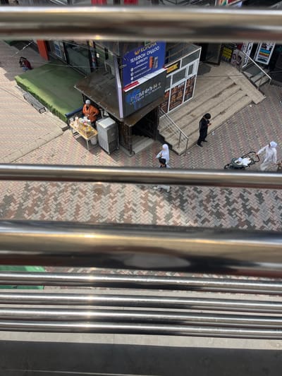 The image shows a street scene in Osh City, Kyrgyzstan, viewed from above. In the foreground, there are several horizontal metal railings reflecting sunlight. Below the railings, the scene unfolds onto a brick-paved sidewalk with a herringbone pattern.

On the left side of the image, a woman in a dark-colored top and red pants is seated on the ground near a green mat. A small bench is visible behind her.

Further into the scene, near a building entrance, a vendor wearing an orange shirt and a light-colored headscarf stands behind a table displaying various items, possibly food or merchandise. To the right of the vendor is a silver-colored refrigerator or freezer unit. Above the vendor, a sign with Cyrillic text and images of cell phones is displayed.

In the middle ground, a set of concrete stairs leads up to an entrance. Several people are visible on or near the stairs. A person in a black tracksuit is standing on the stairs, looking at their phone. Further up the stairs, two people in white head coverings are walking, one of them pushing a baby stroller with a baby inside. Another person in light-colored clothing is walking in the foreground to the left.

The environment appears to be an urban commercial area with buildings, signs, and street furniture. The lighting suggests it is daytime, with bright sunlight casting shadows.