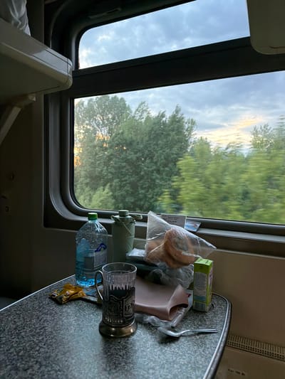 A train window view presents a mixed scene of indoor and outdoor elements. Inside the train, a table is cluttered with various items including a bottle of water, a cup in a metal holder, packaged food, and a spoon. Through the window, a dense green forest is visible under a cloudy sky that transitions to a brighter horizon, suggesting dawn or dusk. The location in the city of Красная Поляна, Russia, cannot be definitively determined from the provided image, as the landscape is not uniquely identifiable.