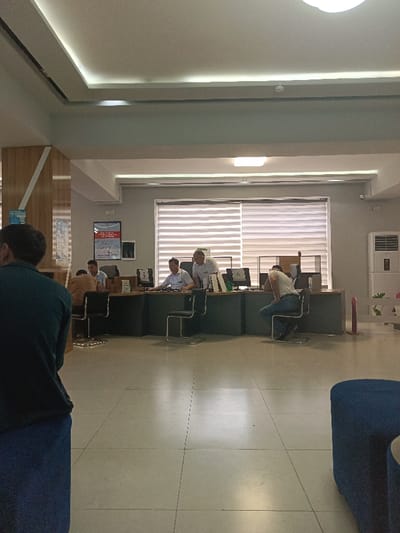 The scene takes place in an office building in Kalgan-Dar'ya, Uzbekistan. The room is a waiting area with a long desk and several chairs. There are four men sitting at the desks, working on computers. The walls are white and there are windows with blinds. The floor is tiled and there is a blue cube-shaped seat near the camera. There is a poster on the wall. The person taking the photo is standing near the blue seat, in the corner of the room. The image is taken from a low angle and the camera is pointed toward the desk. The room is well lit and the people are working quietly.
