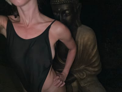 A woman in a black swimsuit is posed next to a dark-faced Buddha statue, with her hand on the statue. The setting is dimly lit and appears to be indoors or in a sheltered outdoor area, possibly in a private residence or a temple setting in Ubud, Indonesia. The woman's skin is visible on her neck, shoulders, and torso. The statue is made of a dark material, possibly stone or resin, with gold-colored drapery. The overall mood is intimate and mysterious due to the low lighting and the juxtaposition of the two figures.