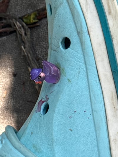 The image displays a close-up view of a light blue plastic surface, possibly part of a playground structure or a garden toy. The surface has a textured pattern and is dotted with small dark holes. In the center of the image, a small, purple, duck-shaped object is attached to the blue surface, seemingly broken off from a larger item. The duck has a distinct head with a beak and an eye. There are some reddish-brown stains and small green smudges on the blue plastic around the object and the holes.
In the background, the ground appears to be covered in a rough, gray material, with some dry, dark-colored twigs or debris visible. The lighting suggests it is daytime, possibly outdoors, with shadows indicating sunlight. The overall impression is that of an outdoor setting in Balivka, Ukraine, where a small toy has become detached and is now a focal point on a playground or garden feature. There is no visible text in the image.