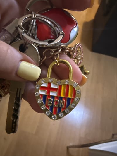 The image shows a close-up of a hand holding a collection of keychains. The keychains are made of metal and enamel, with a heart-shaped charm featuring the colors and crest of FC Barcelona. The crest has a red and yellow striped pattern with a blue and red cross. The letters "FCB" are visible in blue on the crest. The heart-shaped charm is also adorned with small clear crystals. Other keychains include a red apple-shaped charm with a silver casing and a circular silver charm with a hole in the center. A single key is also visible, attached to the keychains. The hand holding the keychains has fingernails painted a pale yellow color. The background appears to be a wooden floor. The scene suggests someone is showing off or organizing their keys. There are no explicit indicators of time of day, weather, or specific emotions, but the focus on the decorative keychains might suggest a sense of personal interest or pride in the team. The location context is Getafe, Spain.