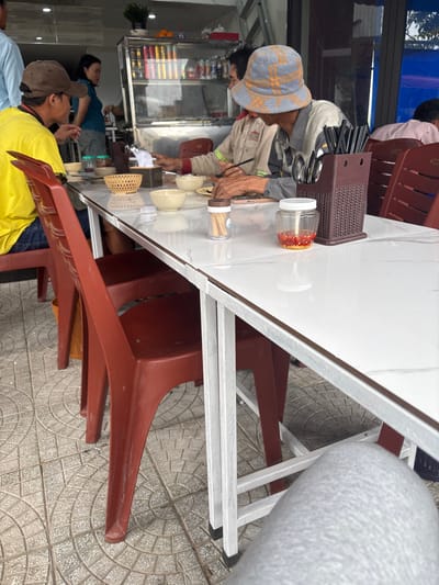 The image depicts a casual open-front eatery in Ngũ Hành Sơn Ward, Vietnam, during daylight. A white rectangular table with a light grey marble-patterned surface occupies the foreground and mid-ground, supported by white metal legs. Four individuals are visible, with the perspective taken from a fifth person whose leg, clad in grey fabric, is seen in the lower right.
On the right side of the table, a male subject wearing a grey and orange checkered bucket hat and a light-colored long-sleeved shirt is seated, actively using black chopsticks to eat from a small, light-colored ceramic bowl. A smartphone is near his right hand. Opposite him, on the left, a male subject in a yellow short-sleeved shirt and a brown baseball cap is partially visible, also seated at the table.
Table accessories include a dark brown plastic holder containing black chopsticks and silver spoons, a clear glass jar with red chilies or chili flakes and a white lid, a white toothpick dispenser with a brown lid, a dark brown tissue box cover with a white tissue, and a light brown woven basket. Several empty small, light-colored ceramic bowls are also present. The chairs are dark red/brown plastic. The floor is covered with grey interlocking paving tiles.
In the background, a female subject in a blue t-shirt stands behind a glass display counter, facing the diners. A green potted plant is on top of this counter. A partial view of a person in a light blue shirt is visible on the far left, and another person in a pink shirt is partially seen at a distant table on the far right.