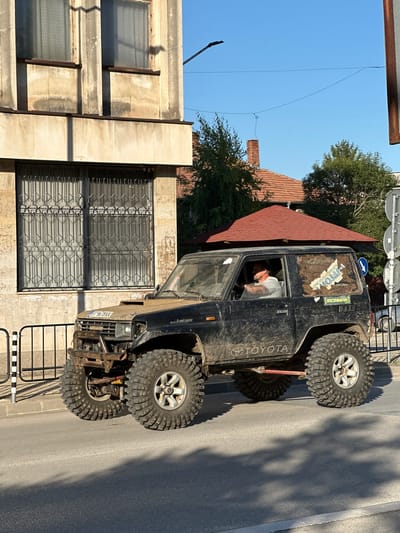 The image captures a modified black Toyota Land Cruiser J80 series, equipped with oversized off-road tires and a reinforced front bumper, parked on a paved street in Lyaskovets, Bulgaria. The vehicle is dirty, suggesting recent off-road use. A middle-aged man with a beard, wearing a grey t-shirt, is visible inside the driver's side, holding the steering wheel. The vehicle has stickers on its rear window, one appearing to be a map of an area with Cyrillic text and another that reads "IRONMAN."

To the left of the vehicle, a building with a beige facade features two large windows with decorative metal grates. The building's architecture suggests it is an older structure, possibly residential or commercial. Sunlight casts shadows from the building and trees onto the street.

In the background, a red-tiled roof of another building is visible, along with green trees and a clear blue sky. Traffic signs and a lamppost are also present. The overall scene depicts a moment in a town street, highlighting the rugged appearance of the modified vehicle against an urban backdrop in Lyaskovets, Bulgaria.