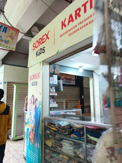 The image depicts an indoor commercial setting in Kidapawan, Philippines, featuring the storefront of a retail establishment. The dominant feature is a yellow overhead sign displaying "SOREX KIDS" in red and black text, with additional text "KARTI" partially visible. Below "KARTI", the Indonesian phrase "MENYEDIAKAN PAKAIAN I PELENGKAPAN BA" is present, translating to "provides clothes and baby accessories." A vertical banner to the left of the entrance features the "SOREX KIDS" logo and an image of two young girls. The store's interior is visible, showing glass display cases filled with folded clothing items. Shelves behind the counter hold various packaged goods, possibly toiletries or baby products. A single white bulb illuminates the store's interior from the ceiling. In the background of the store, two metal rolling shutters are closed. A partial sign with red text "HUB: 08**-**** 8156 NO. ONLY" is visible inside. To the left, a person wearing a yellow and black hooded jacket is partially visible from the back, facing away from the camera. Further left, another commercial unit is visible with its rolling shutter closed. Above and to the left of the main storefront sign, a smaller white sign featuring Winnie the Pooh and text "KA" and "PO. 13-22-53-62" hangs. The floor is tiled, and the ceiling shows exposed concrete beams.