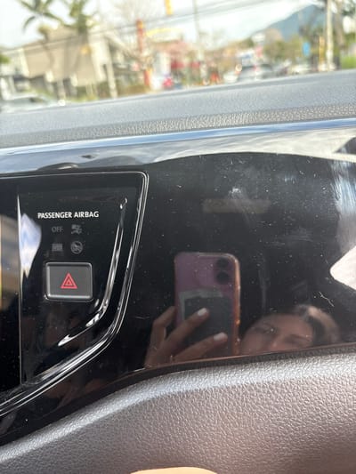 The image shows the passenger-side dashboard of a vehicle. The dashboard is black and has a glossy finish, showing a reflection of a person holding a cell phone. The person's face and hand are visible in the reflection. The dashboard features a passenger airbag control switch, with the "OFF" option selected. The surrounding area is blurry, suggesting the vehicle is in motion. The coordinates (latitude: 9.937, longitude: -84.152) place the location in San Rafael, Costa Rica, but there are no additional clues in the image to confirm the specific location.