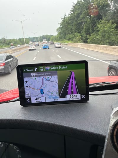 This is a first-person view from the driver's seat of a vehicle, looking at a Garmin GPS device mounted on the dashboard. The GPS displays a map of the area around Danbury, Connecticut, with a route highlighted in purple. The device shows the current speed as 45 mph, and the distance to the destination as 964 meters. The route is on Interstate 84, and the device indicates that the driver is approaching the state border. The background shows a highway with other vehicles, trees, and a cloudy sky. The dashboard of the vehicle is visible in the foreground, including a speedometer with markings up to 95 mph.