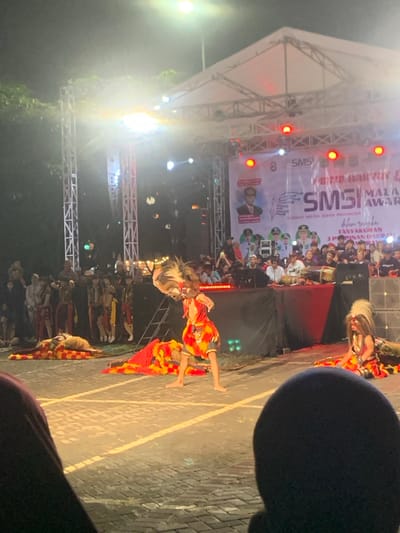 Two people are performing a traditional dance in front of a stage in Kecamatan Batu, Indonesia. One dancer, wearing a yellow and red costume with a large feathered headdress, is in mid-air, kicking their right leg forward. The other dancer, wearing a similar costume with a white headdress, is kneeling on the ground, facing the audience. Both performers have their faces obscured by their masks. The stage is decorated with red and white banners, a large banner displays an inscription in Indonesian. There are many people seated in the audience, most with their backs turned to the camera, Some members of the audience are holding up cellphones, possibly recording the performance. The performance is taking place outdoors on an asphalt or concrete surface, lit by overhead street lamps. It is night time and the lighting is dim. The location, based on the coordinates provided, is a street in the city of Batu, East Java, Indonesia.