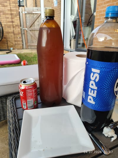 The image captures an outdoor scene at a residence in Wellingborough, United Kingdom, at 9:07 PM on July 4th, 2026. The focus is on a table setting, likely for an evening meal or gathering. On the table, there's a white rectangular plate, a can of Coca-Cola with its top open, a large brown plastic bottle containing a dark liquid, and a bottle of Pepsi. A roll of white paper towels is positioned behind the Coca-Cola can. The table appears to be made of dark woven material, possibly a patio furniture set. In the background, elements of a garden are visible, including a wooden gate, a brick wall, and a patch of grass with a children's toy. The lighting suggests it is dusk or early evening.