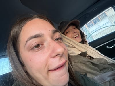The image features two women inside a car during the daytime. The woman in the foreground occupies most of the frame, captured in a close-up, possibly taking a selfie. She has long, dark brown hair, brown eyes, and a fair complexion with freckles. Distinct facial details include a small mole on her forehead, another mole near her nose, a prominent raised blemish on her left cheek, and a small mark or scab on her upper lip. Her mouth is slightly open, suggesting a mild expression or mid-speech.
In the background, seated in the car's passenger seat, is another woman with dark, curly hair, wearing a dark cap, a light-colored striped scarf, and a brown or khaki-colored jacket. She is smiling broadly and looking towards the foreground, indicating a happy demeanor.
The interior of the car is visible, including the dark headliner, seat upholstery, and part of a door panel with a handle. Through the side window, out-of-focus details of buildings with horizontal features are discernible, suggesting an urban or built-up environment. The overall lighting is bright, consistent with daylight. No text is visible in the image. While the location context of Mitzpe Ramon, Israel, is provided, there are no specific visual cues within the image to confirm this.