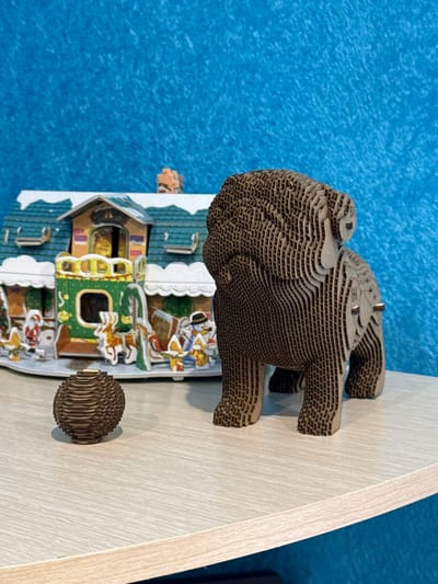 A cardboard Christmas-themed diorama sits on a light-colored wooden surface. The diorama depicts a house decorated for Christmas with cartoonish figures and green and yellow trim. Perched atop the house is a layered cardboard animal figure, possibly a bear, and to the right is a small, spherical cardboard object. The background consists of a blue, textured surface. The scene is in Chuprino, Russia.