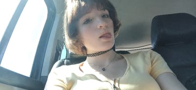 The image depicts a single individual, a person with short, light brown hair with bangs, light skin, and freckles, seated within a vehicle. The person is looking directly forward, with their tongue slightly extended and resting on their lower lip. A small septum piercing is visible. They are wearing a light yellow top, a black lace choker, and a silver-colored chain necklace with a dark pendant. To the individual's left, a vehicle window is visible, showing a bright, overexposed sky, suggesting an outdoor, daytime setting. A dark headrest is visible behind and to the individual's right. The location is specified as the city of Bistrice, Montenegro.