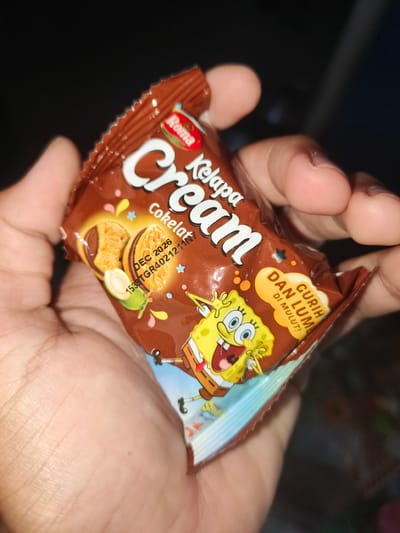 A person's hand is holding a chocolate cookie package featuring SpongeBob SquarePants. The package is brown with white lettering that says "Roma Cream Kelapa Cokelat". There are also images of cookies and a halved coconut on the package. The person's thumb and forefinger are visible on the right side of the frame, holding the package. The background is dark and out of focus, with some hints of what might be a dimly lit indoor environment. The image appears to be taken in Barengkrajan, Indonesia, as suggested by the presence of the product.