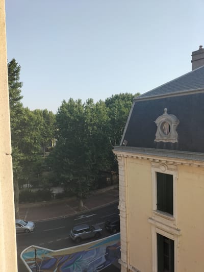 The image shows an urban scene from an elevated perspective, likely a window or balcony, looking out over a street and adjacent buildings. The setting appears to be a city, with trees, a road, and architecture.
Specifically, the scene is located in Narbonne, France, as indicated by a previous analysis.
In the foreground, a textured, light-colored wall or pillar is visible on the left side, partially obscuring the view. Below and slightly to the right of this pillar, a street is visible with two cars. A silver sedan is parked on the left side of the road, and a silver SUV is driving on the right side of the road. There is also a black hatchback parked on the right side of the road. The road has white lane markings.
Beyond the road, there is a green area with numerous trees, suggesting a park or tree-lined boulevard. The foliage is dense and lush, indicating a temperate climate, possibly summer or late spring.
On the right side of the image, a building with a pale yellow facade and a dark gray, mansard-style roof is prominent. The building features decorative stonework around its windows and along the roofline. A distinctive oval dormer window with decorative trim is visible on the roof. Two tall, dark-shuttered windows are present on the facade of this building.
The sky above is a clear, pale blue, suggesting daytime, likely in the morning or late afternoon due to the angle of the light. The overall impression is one of a quiet, residential or commercial street in a European city.