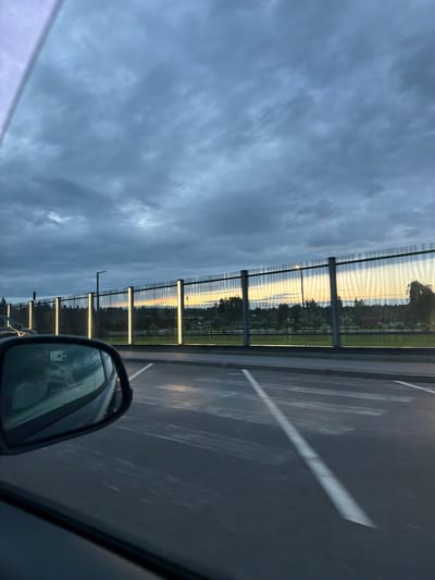 The image shows a cloudy sky over a cityscape in Solnechnoye, Russia. A reflective fence with vertical lights separates a road from trees and buildings in the background. A car's side mirror is visible in the foreground, reflecting a person and part of the car's interior. The road has white lane markings and appears to be a parking lot or an area with parking spaces. The lighting suggests it is either dawn or dusk.