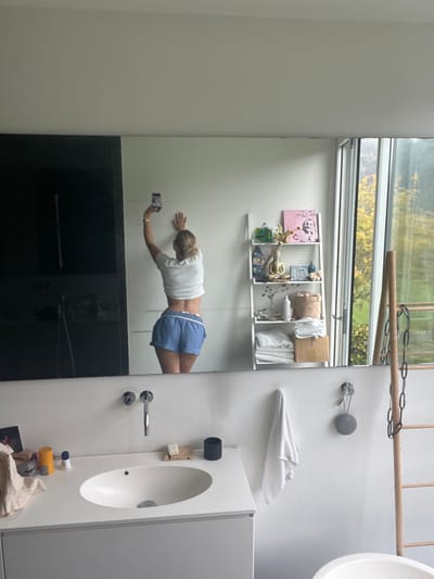 An adult individual, with blonde hair pulled back, is reflected in a large rectangular mirror in a modern bathroom. The individual, dressed in a white cropped t-shirt exposing the midriff and blue shorts with white trim, is seen from a rear-right angle, extending their left arm upward to hold a smartphone, capturing a mirror selfie. The right hand rests against the white wall behind them.
The bathroom features a white vanity with a single oval sink and a chrome wall-mounted faucet. The white countertop holds various items including toiletries, a bar of soap on a wooden tray, and a light-colored cloth. To the right of the sink, a white towel hangs on a chrome hook, with a grey circular smart speaker mounted above it. Further right, a wooden ladder-like structure with dark chains is visible. The edge of a white freestanding bathtub is partially visible in the bottom right foreground.
Reflected on the wall behind the individual is a white open shelving unit displaying decorative items such as a golden Buddha statue, various containers, and a pink square art piece. Folded white towels and a cardboard box are on the lower shelves. To the right of the shelving unit, a large vertical window reflects an outdoor scene with green and yellow-leafed trees under an overcast sky, indicating an exterior view consistent with a residential area.
The scene is located within the city of Skanderborg, Denmark.