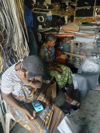 The image displays an interior view of a commercial or workshop space in Kwekwe, Zimbabwe. Two individuals are seated, surrounded by various materials and merchandise. In the foreground, a person with dark, wavy braided hair, wearing a horizontal striped t-shirt over a patterned brown, orange, and white skirt, and white trousers, is seated on a white plastic chair. They are looking down at a light blue electronic device, possibly a point-of-sale terminal or calculator, held in their lap. Their right hand is near their right ear/temple. A yellow bracelet is visible on their right wrist.
In the midground, a second individual with braided hair, dressed in a green and yellow patterned dress, is seated on a wooden chair, also looking downwards. Their right foot, wearing a light-colored slide sandal, is visible. To the far left, numerous bundles of silver and gold-toned synthetic braiding hair or cords hang vertically. The background consists of wooden shelving units densely packed with stacks of fabric or leather sheets in various colors (grey, brown, black), rolls of material, small clear jars with lids, and large white sacks containing bulk goods. A brown cardboard box with an upward arrow symbol is present on the concrete floor. A third person, wearing a blue shirt, is partially visible in the left background behind the hanging materials.