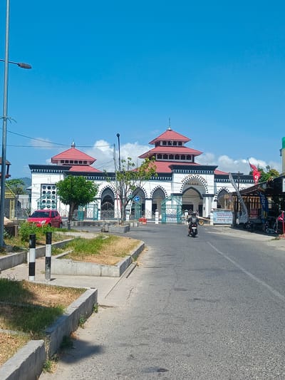 This is an eye-level, full shot image of a mosque in Luwuk, Indonesia. The mosque has white walls with black trim, and red tiered roofs. A white sign above the arched entrance reads "MASJID AR-RAHMAN LUWUK". In front of the mosque, there is a street with a single motorcycle rider in the foreground, heading away from the camera. To the left of the street, there is a red car parked next to a patch of dry grass and concrete planters. A street lamp is visible on the left side of the image. The sky is bright blue with scattered white clouds.