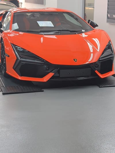 A bright orange sports car, possibly a Lamborghini, is the main subject of this image. It is viewed from a low angle, tilted to the right. The car is parked indoors, on a grey, textured floor, with a light-colored wall behind it. Part of the car's interior, including seats and a reflection on the windshield, is visible. The car has sharp, angular lines and black accents. A black plate holder is mounted on the front of the car. The scene appears to be a showroom or garage. There is no text visible in the image. The lighting suggests an indoor environment, possibly during the day.