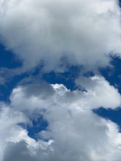 A flock of birds flies in a V-formation against a bright blue sky filled with white and gray clouds. The birds are small and appear as dark specks in the distance. The clouds are scattered and fluffy, with some areas of shadow indicating varying thickness and density. The image captures a natural outdoor scene.