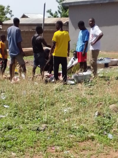 The scene depicts a group of approximately seven people gathered in an open, grassy area. The setting appears to be outside in daylight, possibly in the vicinity of Dungu, Ghana. Buildings with corrugated roofs are visible in the background. The ground is covered with a mixture of tall grass and what appears to be scattered debris. The group is clustered around several metal basins and some kind of meat, which appears to be being processed. The people are wearing various casual clothing items, including shirts, shorts, and trousers.