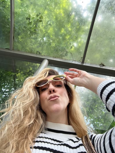 A woman with long blonde curly hair is adjusting yellow-framed sunglasses. She is wearing a white and black striped sweater and a metallic belt. She is in a glass structure, possibly a greenhouse or bus stop, with green foliage visible through the glass. The location may be Dronten, Netherlands.