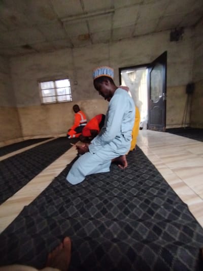 The image shows three men kneeling on a black and white patterned prayer rug in a room in Ashaka, Nigeria. The man in the foreground is wearing a blue and yellow robe and a white cap, and he is looking down at his phone. Two other men are kneeling behind him. The room is sparsely decorated, with a single window and a door leading to the outside. The door is open and a glimpse of foliage can be seen. The floor is tiled. The men are likely praying, but this cannot be confirmed from the image.